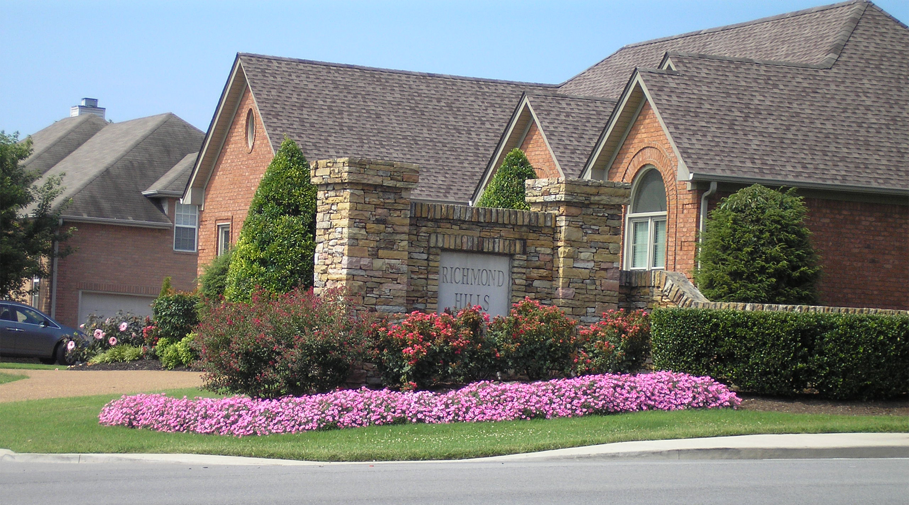 Picture of a stone neighborhood sign surrounded by pink flowers and red bushes.